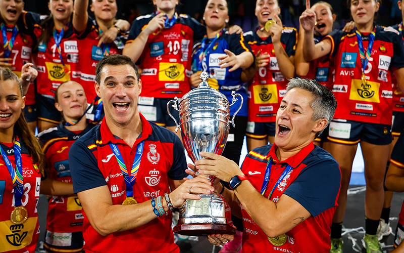 A group of athletes in red and blue uniforms celebrate with a large silver trophy, smiling and cheering while wearing medals.